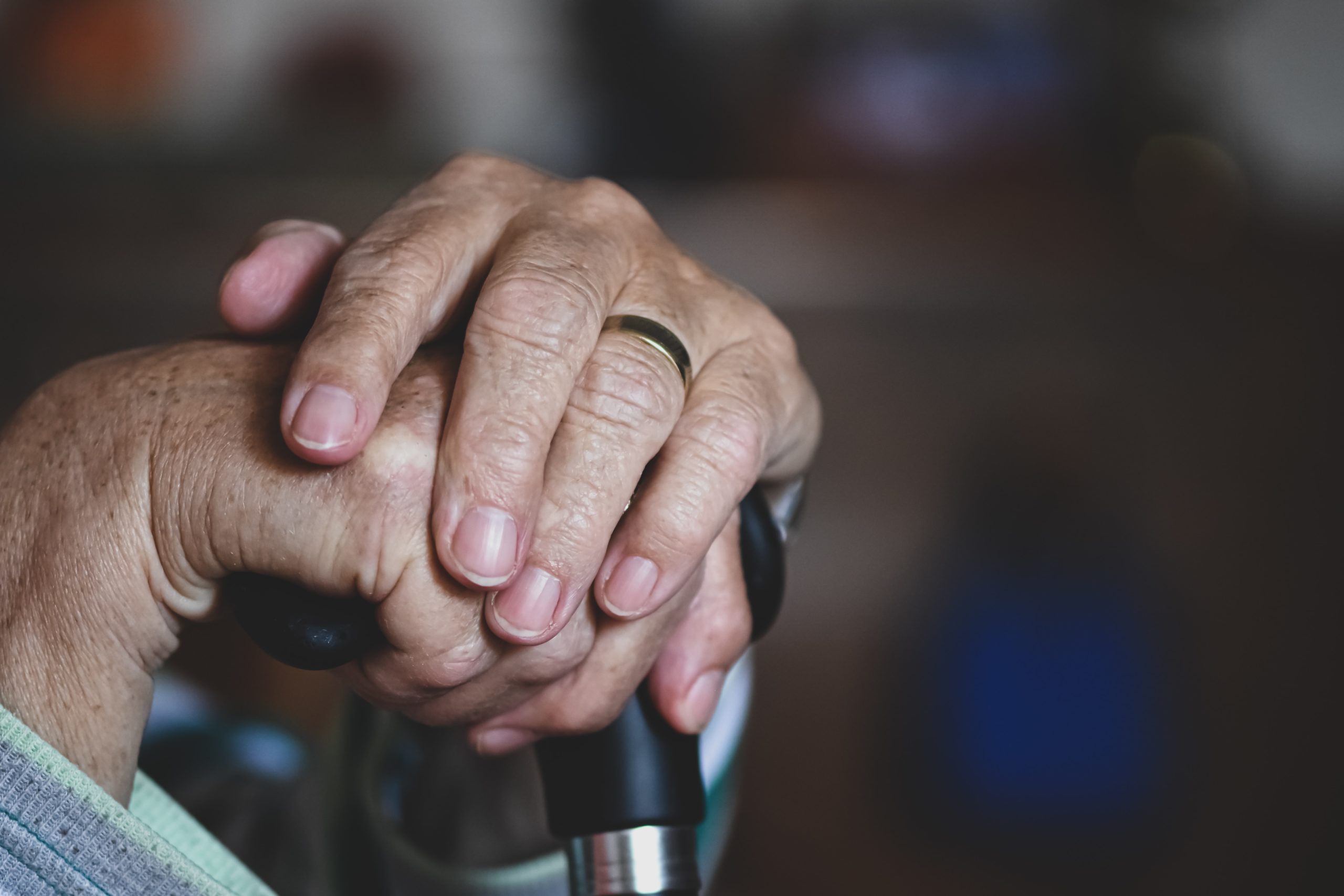 Hands of a senior citizen with a wedding band on ring finger, resting on a cane.