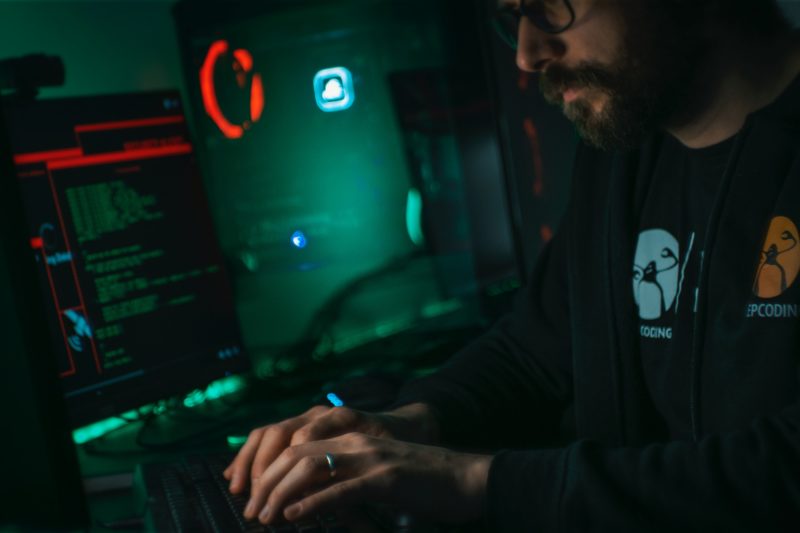 Man in sweater, typing on monitor computer at a desk, in a dark, dimly lit room.