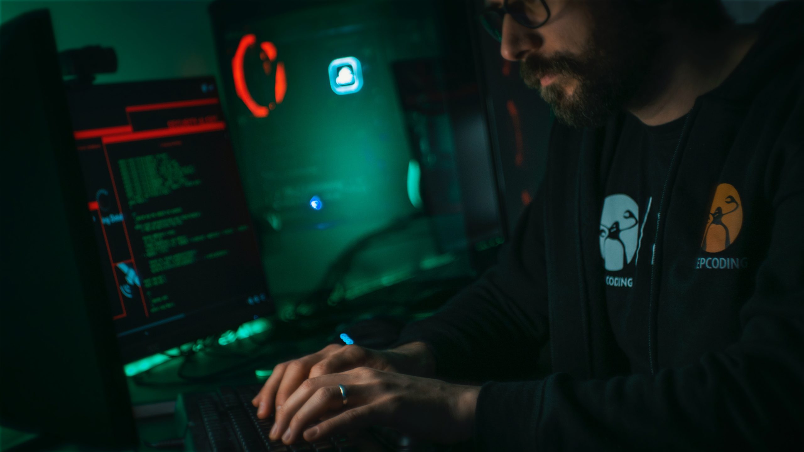 Man in sweater, typing on monitor computer at a desk, in a dark, dimly lit room.